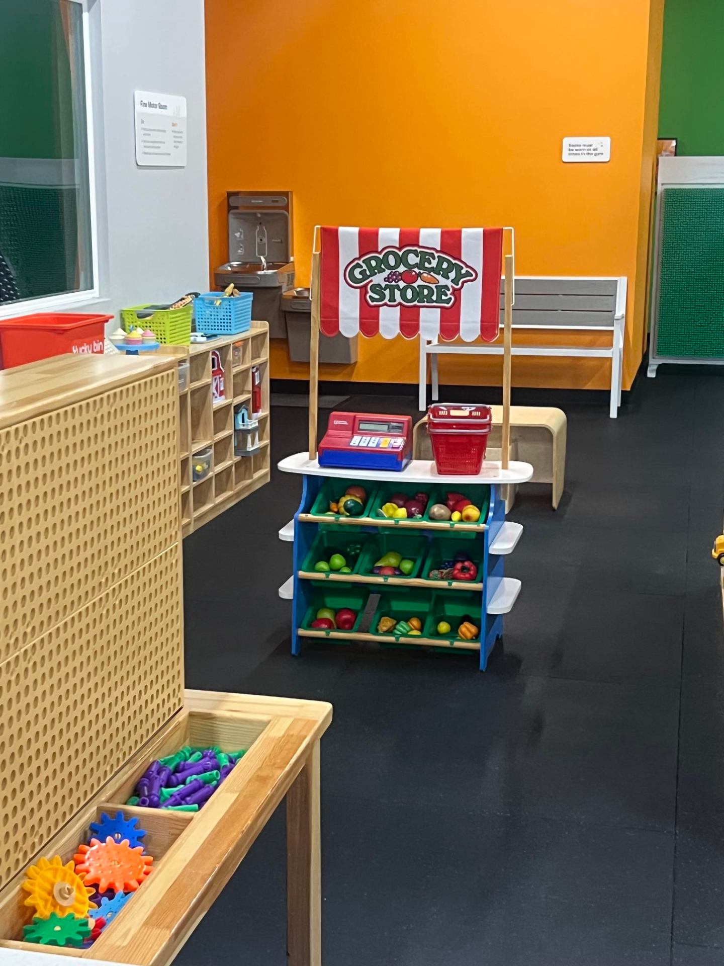 Pretend grocery store play area with produce bins, register, and bright orange wall at Little Land New Braunfels