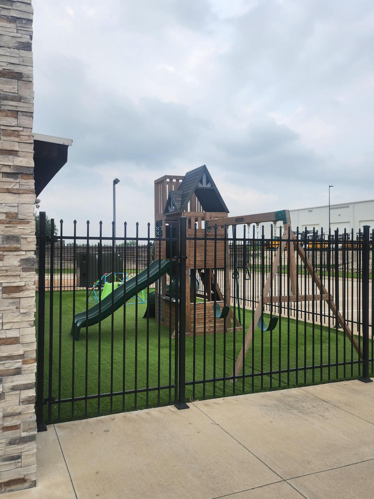 Fenced outdoor playground with wooden play structure, green slide, and swing set at Little Land Buda