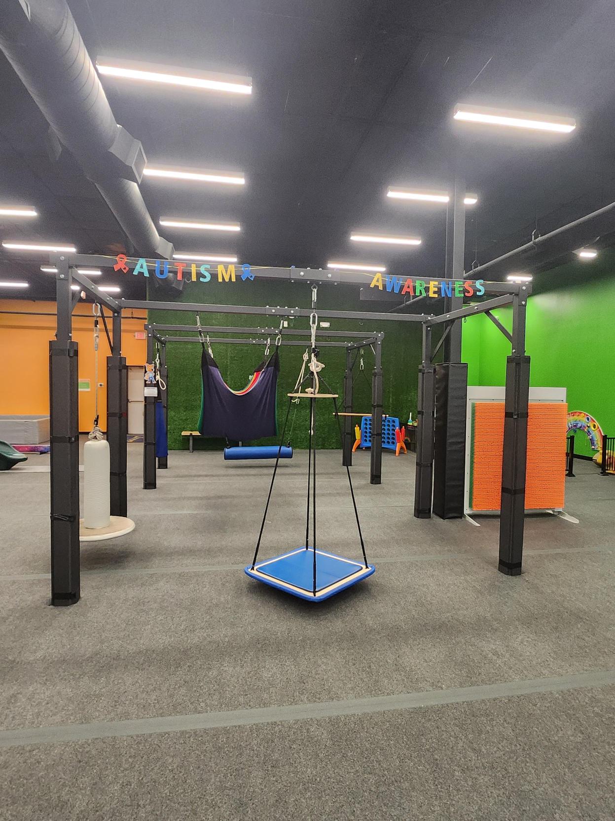 Sensory swing therapy gym with platform swing, hammock, and climbing equipment under an Autism Awareness banner