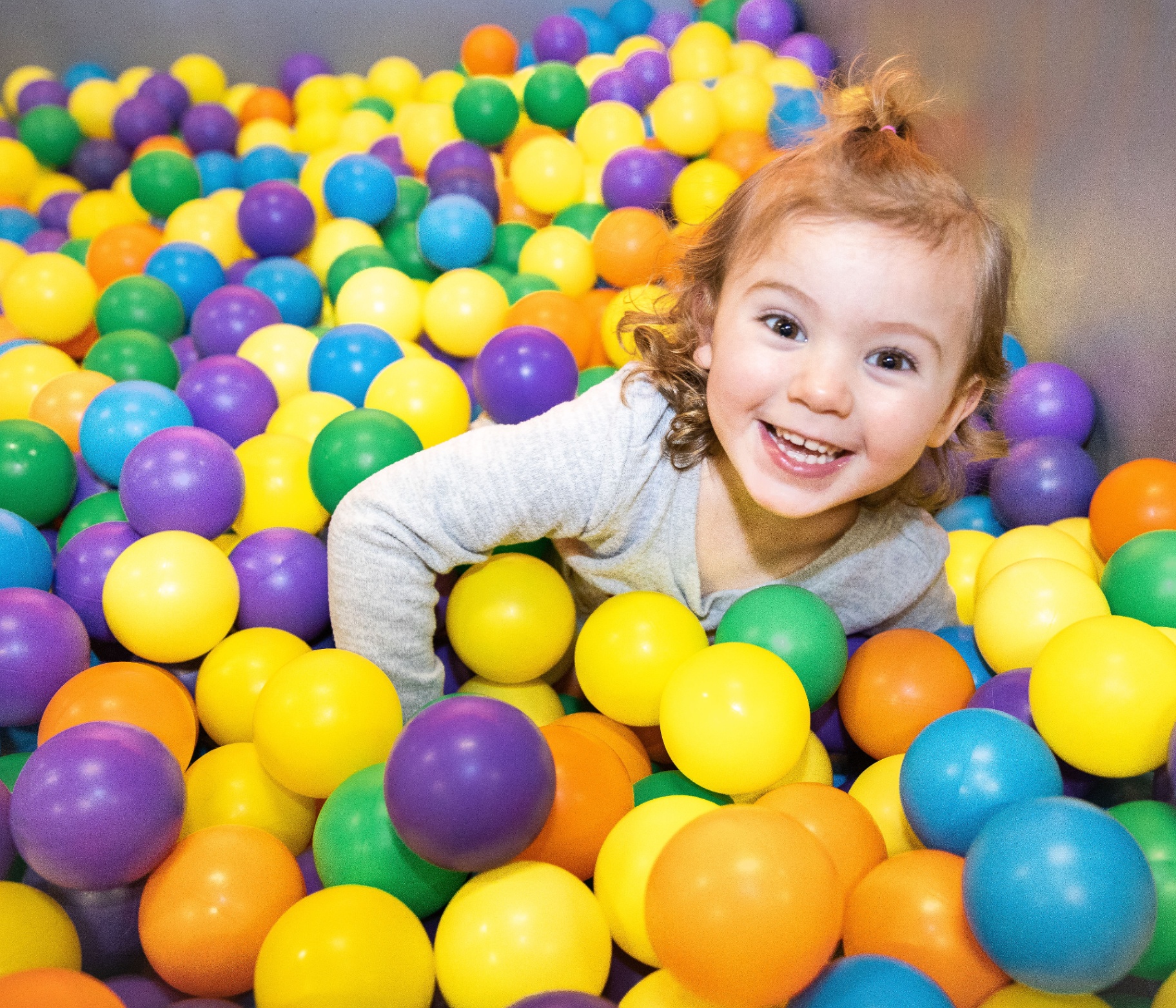 Toddler beaming inside a rainbow ball pit
