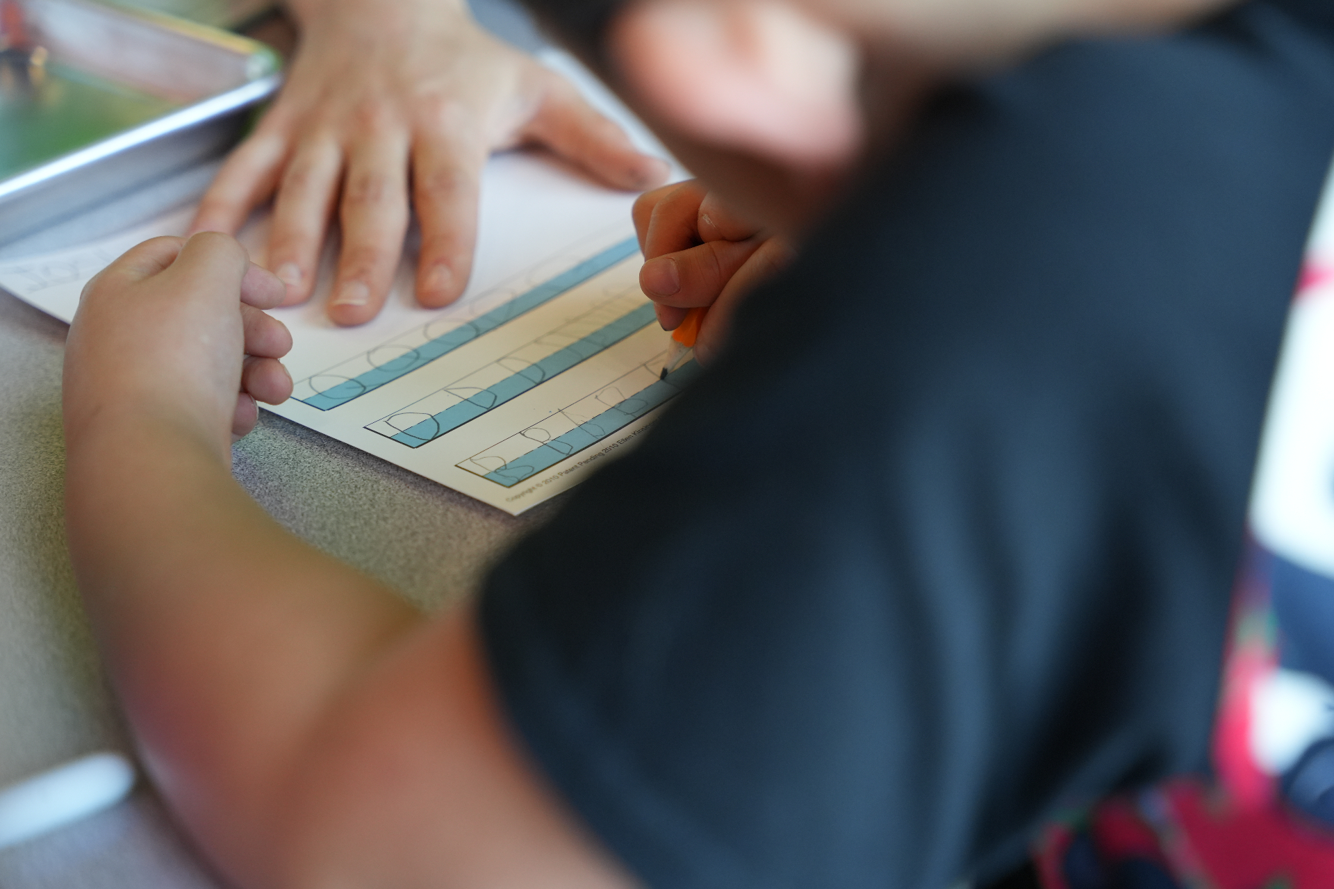 Close-up of a child practicing letter tracing with a teacher's hand guiding the page